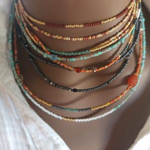 May include: A selection of layered beaded necklaces in shades of red, orange, turquoise, and black. The necklaces are displayed against a white collared shirt. The small beads create a delicate, bohemian style. The necklaces are of varying lengths.