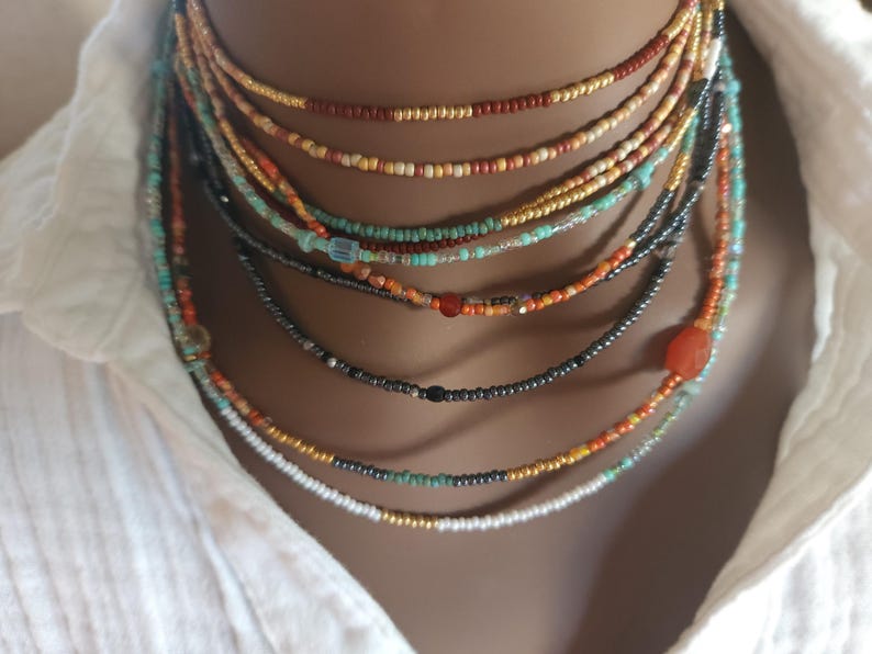 May include: A selection of layered beaded necklaces in shades of red, orange, turquoise, and black. The necklaces are displayed against a white collared shirt. The small beads create a delicate, bohemian style. The necklaces are of varying lengths.