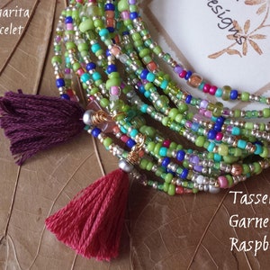 May include: A multi-strand beaded bracelet with green, blue, silver, and pink beads. The bracelet has two tassels, one burgundy and one red. The text "Margarita Bracelet" and "Tassels: Garnet Raspberry" is visible in the image.