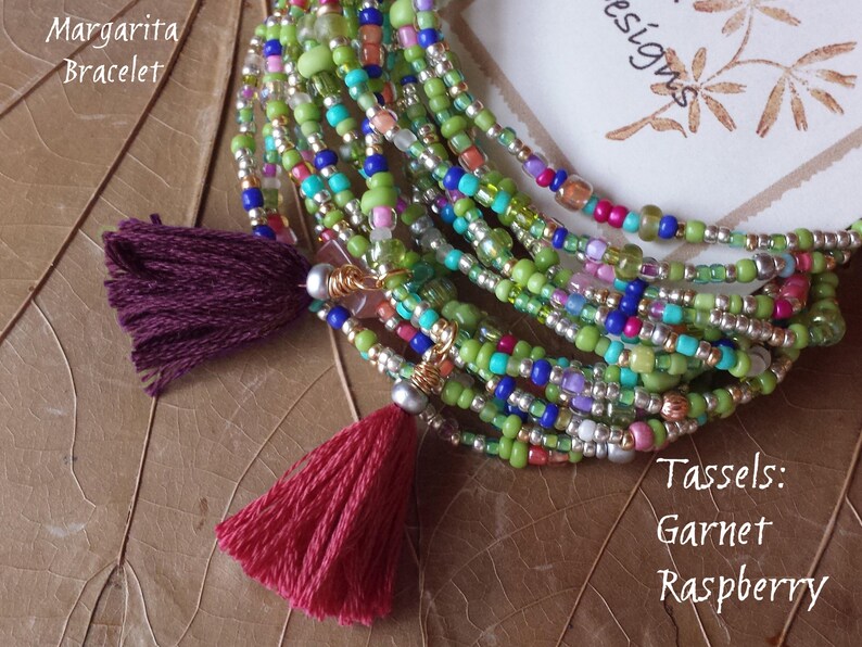 May include: A multi-strand beaded bracelet with green, blue, silver, and pink beads. The bracelet has two tassels, one burgundy and one red. The text "Margarita Bracelet" and "Tassels: Garnet Raspberry" is visible in the image.