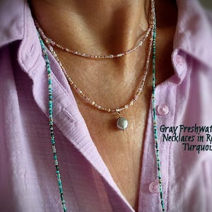 May include: Three layered necklaces with a gray freshwater pearl pendant. The necklaces are rose and turquoise in colour. The image also shows a light purple button-down shirt.