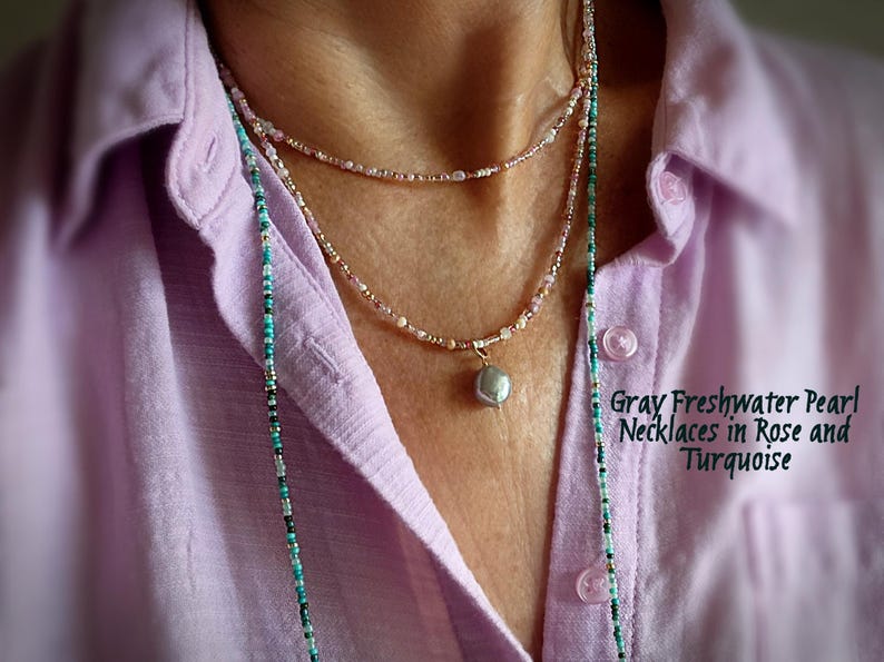 May include: Three layered necklaces with a gray freshwater pearl pendant. The necklaces are rose and turquoise in colour. The image also shows a light purple button-down shirt.