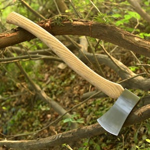 May include: A wood-handled axe with a silver blade. The axe handle is light brown and has a curved design. The axe head is silver and has a sharp edge. The axe is resting on a tree branch.