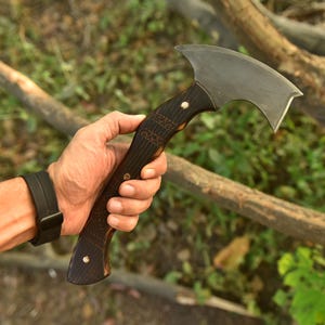 Puede incluir: Un hacha artesanal con un mango de madera oscura y una hoja curva de color plateado. El mango presenta intrincados detalles tallados y se sostiene en una mano sobre un fondo natural borroso. El hacha parece ser una herramienta o un art&iacute;culo decorativo.