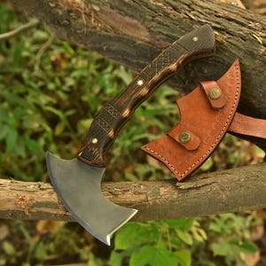 Puede incluir: Un hacha peque&ntilde;a y curvada con un mango de madera oscura y una funda de cuero. La cabeza del hacha es plateada y el mango presenta tallas decorativas y remaches de lat&oacute;n. La funda de cuero es marr&oacute;n con cierres de bot&oacute;n de lat&oacute;n.
