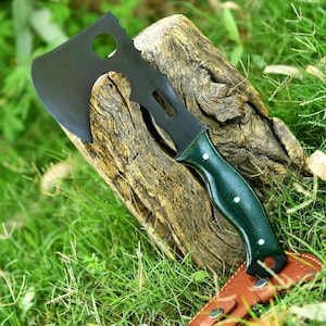 May include: A black hatchet with a green handle and a brown leather sheath rests on a weathered wooden log. The hatchet has a curved blade with a hole and a serrated edge. The handle is dark green with three white dots.