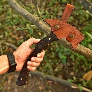 Puede incluir: Un hacha artesanal con un mango de madera oscura y una funda de cuero marr&oacute;n. La cabeza del hacha es de metal y tiene una hoja curva. La funda de cuero se asegura con dos remaches color lat&oacute;n. El mango tiene un dise&ntilde;o texturizado.