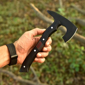 May include: A black hatchet with a curved blade and a wooden handle. The handle is dark with white dot accents and a red-brown inlay. The hatchet is held in a hand against a blurred green and brown background.