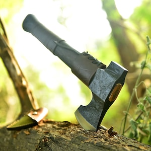 May include: A forged steel axe with a dark wooden handle and a leather sheath. The axe head is a dark gray color with a bronze accent. The axe is resting on a log in a forest setting.