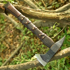Könnte beinhalten: Eine handgefertigte Axt mit einem dunklen Holzgriff, der mit braunen Lederriemen umwickelt und mit Messingnieten befestigt ist. Der Axtkopf besteht aus dunklem Metall mit einer scharfen, gebogenen Klinge. Die Axt liegt auf einem Ast.