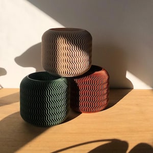 Könnte beinhalten: Drei runde, dekorative Pflanzer aus recyceltem Papier. Die Pflanzer sind übereinander gestapelt. Der obere Pflanzer ist beige, der mittlere Pflanzer ist grün und der untere Pflanzer ist orange.