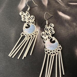 May include: A pair of silver-toned dangle earrings. Each earring features a peacock design with a blue and silver-toned body and a decorative feather design. Dangling from the peacock are several silver-toned rods.