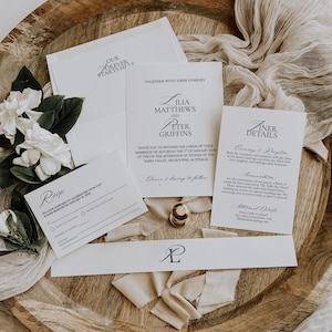 May include: A stylish wedding invitation suite featuring white cards, envelopes, and RSVP cards. The suite includes a gold wax seal, a ribbon, and white flowers. The invitation text reads "Ilia Matthews and Peter Griffins".