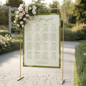 May include: A rectangular wedding seating chart sign with a green border and white background, featuring the text Find Your Seat and Camilla & Antonio 01.01.2030. The sign is displayed on a gold metal stand adorned with white roses and green foliage.