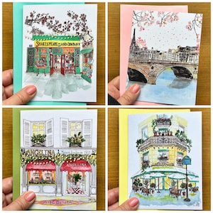 Könnte beinhalten: Vier Aquarell-Illustrationen auf Grußkarten. Die erste Karte zeigt ein grünes Gebäude mit dem Text "Shakespeare and Company". Die zweite Karte zeigt eine Brücke über einen Fluss mit einem rosa Himmel und fallenden weißen Blüten. Die dritte Karte zeigt ein gelbes Gebäude mit dem Text "Angelina" und den Worten "Maison fondée en 1903". Die vierte Karte zeigt ein gelbes Gebäude mit einem grünen Vordach und dem Text "Angelina".
