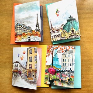 May include: Set of six greeting cards featuring watercolour illustrations of Parisian landmarks, including the Eiffel Tower, a cafe, and a building with a green dome. The cards are printed on white cardstock and have a blank inside for writing.