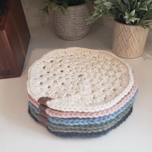May include: A stack of six crocheted coasters in various colors, including white, pink, blue, green, and gray. The coasters are round and have a simple design. The top coaster is white and has a brown leather strap.