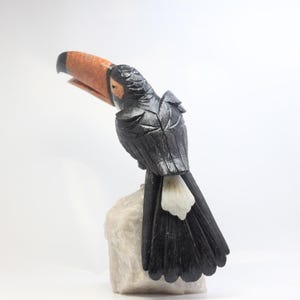 May include: A carved toucan sculpture with a large orange and black beak, perched on a white stone base. The bird's body is black with detailed feather carvings. The sculpture is set against a white background.