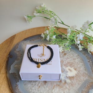 Peut inclure: Un bracelet de perles noires avec un fermoir et un charme dorés repose sur une boîte blanche. La boîte porte le mot "PRISME". Le bracelet est présenté sur une surface décorative avec des fleurs blanches en arrière-plan.
