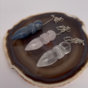 May include: Three crystal pendulums in blue, pink, and clear quartz, each with a silver chain. The pendulums are displayed on a brown and white agate slice. These are ideal for divination and spiritual practices.