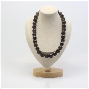 Peut inclure: Un collier de perles rondes brun foncé exposé sur un buste crème. Les perles sont de taille uniforme et enfilées ensemble, créant un aspect lisse et élégant. Le buste est posé sur une base en bois sur fond blanc.