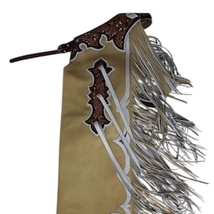 Peut inclure: Chaps en cuir couleur tan avec des broderies blanches et brunes et des franges. Les chaps ont une boucle de ceinture et une sangle.
