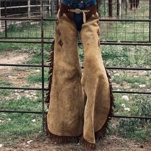 May include: Brown leather chaps with fringe and diamond-shaped stitching. The chaps are worn over jeans and a belt.