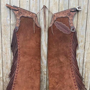 May include: Brown leather chaps with detailed floral tooling on the upper section. The chaps have long fringe along the sides and decorative stitching. A silver buckle and ring are visible.