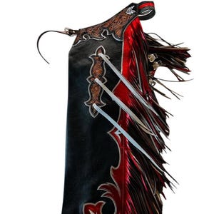 Peut inclure: Chaps en cuir noir et rouge avec des coutures décoratives et des franges. Les chaps présentent un design détaillé avec des accents marron et argent. La frange est un mélange de rouge et de marron.