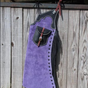 May include: A purple suede chaps with black leather accents and fringe. The chaps feature a black pocket with a decorative strap and a white stitched design. The chaps are hanging on a wooden fence.