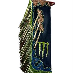 Könnte beinhalten: Ein Paar Lederchaps in Schwarz mit einem hellbraunen, floral geprägten Lederjoch, langen metallisch-limettengrünen Fransen und einem leuchtend grünen Monster Energy-Logo. Die Kanten sind mit blauen Ziernähten und drei hellbraunen Lederbändern versehen.