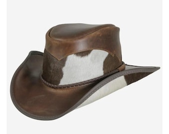 Op de afbeelding: Een bruine leren cowboyhoed met een wit en bruin koeienhuidband. De hoed heeft een brede rand en een hoge kroon.
