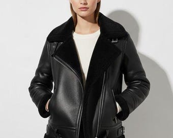 May include: Black faux leather jacket with a shearling collar and lining. The jacket features a diagonal zipper closure, multiple buckles, and a classic aviator style. The jacket has a warm, cozy appearance.