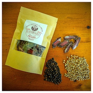 May include: A brown paper bag with a label that reads "Kambako Biltong Original" and a picture of an elephant. The bag contains dried meat. There are also piles of black peppercorns and white peppercorns on a wooden surface.