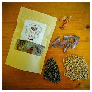May include: A brown paper bag with a clear window showing dried meat inside. The bag has a label that says "Kambako Biltong Spicy" and features an elephant logo. The bag is surrounded by red chili peppers, black peppercorns, and white peppercorns.