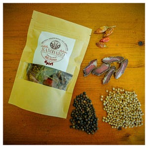 Może przedstawiać: Brązowa papierowa torba z etykietą z napisem "Kambako Biltong Spicy" i suszoną wołowiną. Torba leży na drewnianej powierzchni z czarnymi ziarnami pieprzu, białymi ziarnami pieprzu i czerwonymi papryczkami chili.