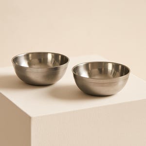 Puede incluir: Dos cuencos pequeños de metal cepillado con un diseño simple y minimalista. Cada cuenco tiene una forma redondeada con un borde ligeramente ensanchado y una sola ranura horizontal cerca de la base. Los cuencos están sobre un fondo neutro.