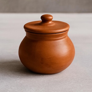Puede incluir: Un pequeño tarro redondo de cerámica de color terracota con tapa a juego. La tapa tiene un pequeño pomo redondeado para facilitar su manejo. El tarro tiene un acabado liso y sin esmaltar.