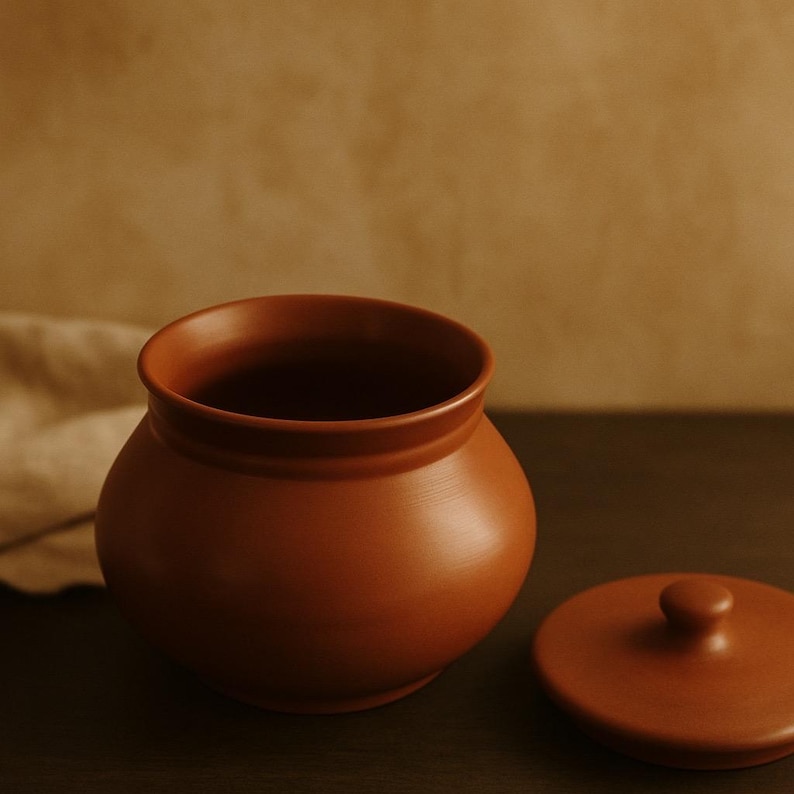 May include: A terracotta-colored ceramic jar with a matching lid. The jar has a rounded body and a wide opening. The lid is round with a small knob on top. The jar and lid are sitting on a dark surface.
