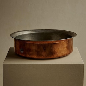 Puede incluir: Un cuenco redondo de metal color cobre con un borde ancho y un interior oscuro. El cuenco está sobre un pedestal cuadrado de tono neutro. Un pequeño número "3" está estampado en el lateral del cuenco.