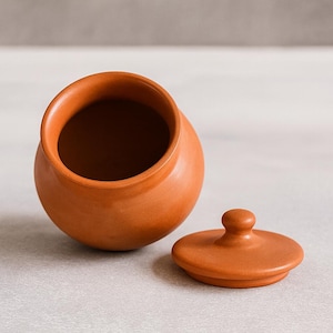 May include: A small, terracotta-colored ceramic jar with a matching lid. The jar is round with a wide opening, and the lid is flat with a small knob. The lid is placed next to the jar.