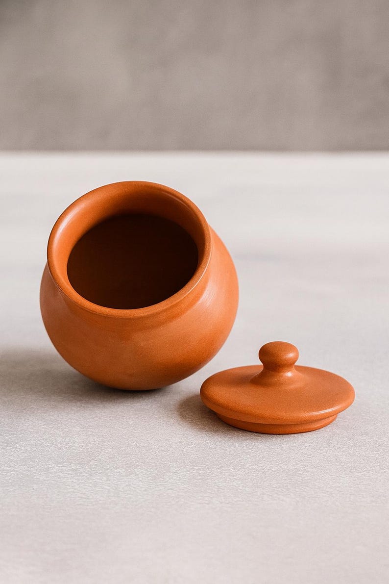 May include: A small, terracotta-colored ceramic jar with a matching lid. The jar is round with a wide opening, and the lid is flat with a small knob. The lid is placed next to the jar.