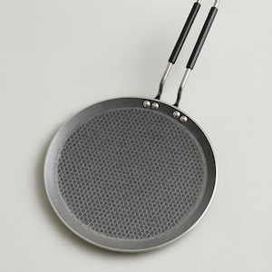 Puede incluir: Una sartén redonda para crepes, de color gris oscuro, con una superficie de cocción con textura de panal. El mango plateado tiene agarres negros. Diseñada para cocinar crepes y otros panqueques finos.