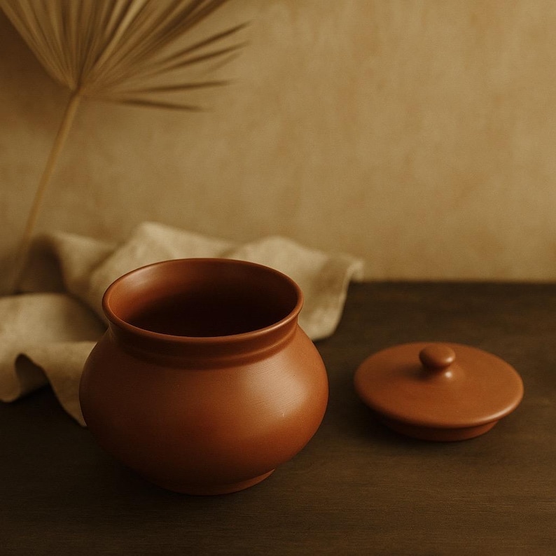 May include: A terracotta-colored ceramic jar with a matching lid. The jar has a rounded body and a wide opening. The lid is circular with a small knob. The jar and lid are on a dark wooden surface.