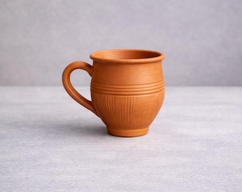 May include: A handmade terracotta mug with a rounded base and a curved handle. The mug has horizontal bands around the middle and vertical lines near the bottom. The mug is a warm, earthy brown color.