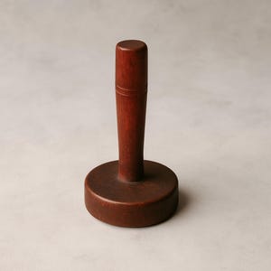 May include: A wooden kitchen tool with a dark reddish-brown finish. It features a cylindrical handle with a decorative groove near the top, connected to a flat, circular base. The tool is designed for food preparation.