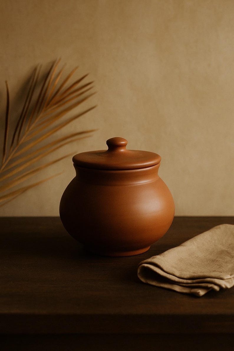 May include: A terracotta-colored ceramic jar with a lid, set on a dark wooden surface. The jar has a rounded body and a small knob on the lid. A folded tan linen napkin rests nearby. The background is a neutral, warm tone.