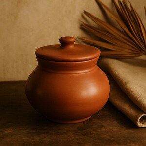 May include: A terracotta-colored ceramic jar with a lid. The jar has a rounded body and a small knob on top of the lid. The background features a neutral-toned wall and a dried palm leaf.