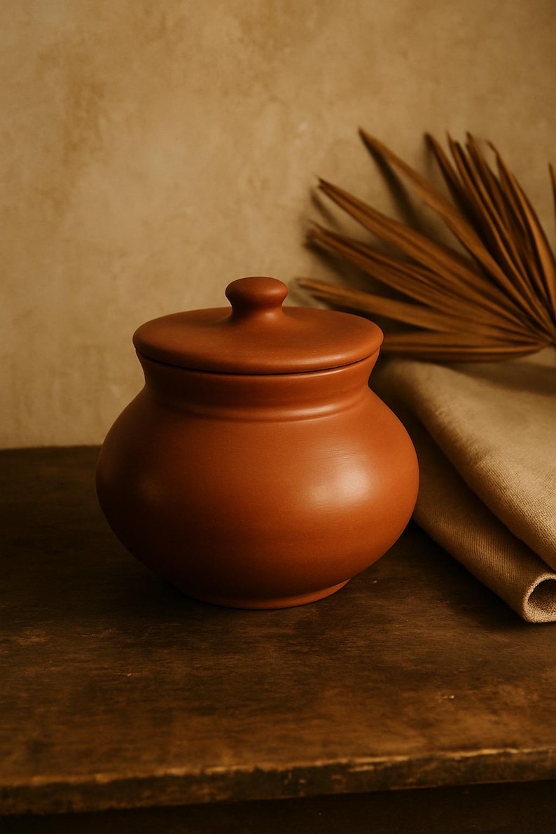 May include: A terracotta-colored ceramic jar with a lid. The jar has a rounded body and a small knob on top of the lid. The background features a neutral-toned wall and a dried palm leaf.