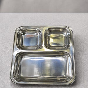May include: A stainless steel divided plate with three compartments. The plate has a rectangular shape with rounded corners and a polished, reflective surface. Two smaller square compartments are at the top, and a larger rectangular compartment is below.